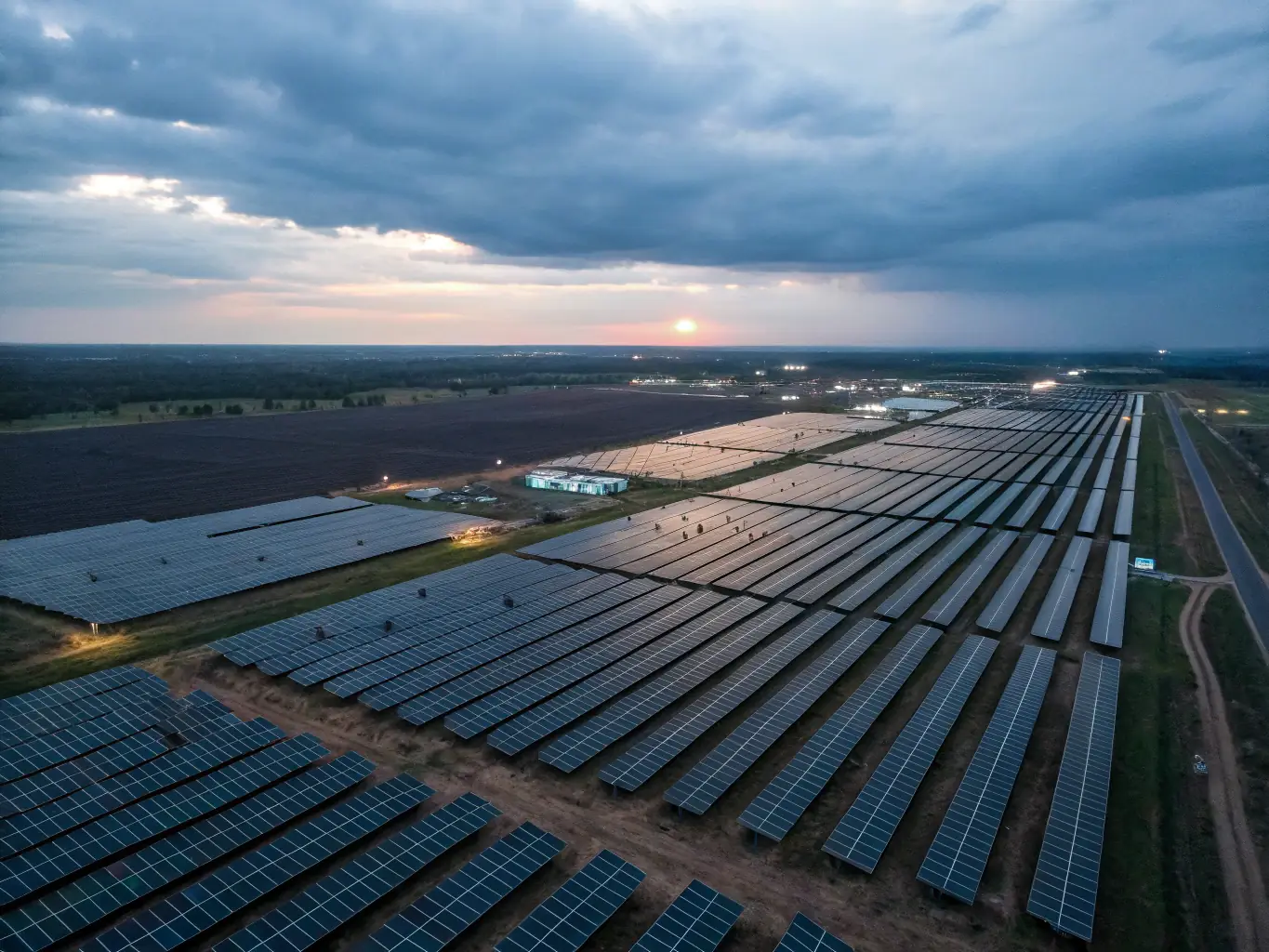 A high-angle, wide shot of a modern, grid-scale battery power plant at sunset, showcasing rows of sleek, silver battery containers with solar panels in the background, set against a vibrant sky.